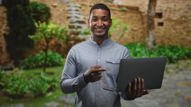 Man pointing finger at laptop while holding it with his other hand in a building courtyard with stone wall and plants; confidence professionalism.