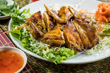 A white plate displays golden grilled chicken pieces on shredded cabbage, garnished with carrot flower and served with chili sauce.