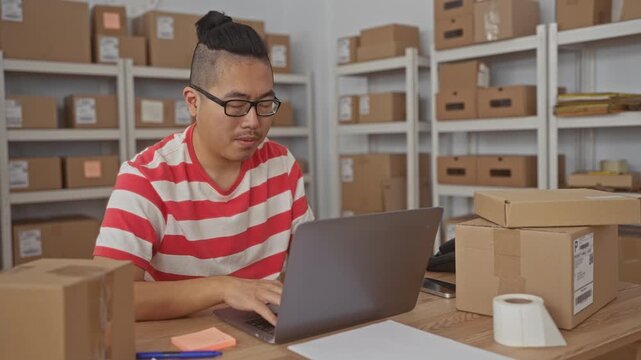 Man typing on laptop and points finger to laptop inside building filled with stacked parcels and shelves while checking labels and tape; focus productivity.
