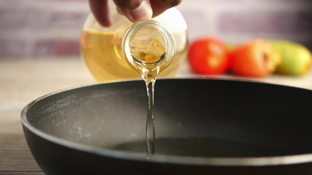 Cooking oil pouring slowly from glass bottle into black frying pan for preparing food
