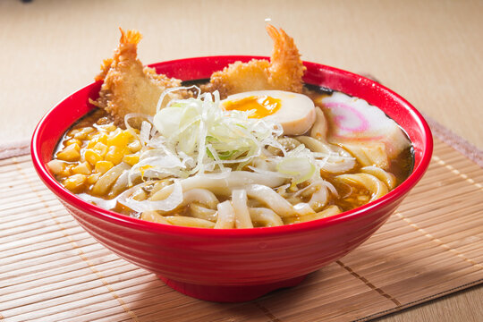 A red bowl of thick udon noodles in curry broth topped with fried shrimp, boiled egg, corn, and sliced vegetables.