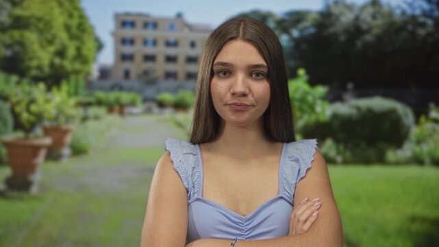 Girl crosses arms in green park near building with potted plants and stone pathway under sunny daylight; denial.