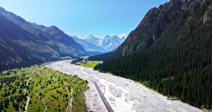 Aerial view of a winding highway through lush green valleys and snow-capped Tianshan Mountains in Xinjiang, China. Road trip landscape.