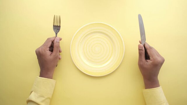 Top view of person holding fork and knife over empty yellow plate on yellow background