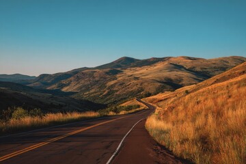 Winding asphalt road curves through golden fields toward rolling mountains under clear sky