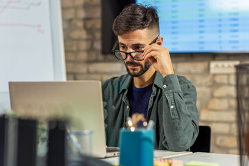 Obraz premium Close-up of serious young man adjusting glasses while using a laptop in modern office.