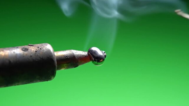 Hot soldering iron tip melting solder wire with smoke on green background.