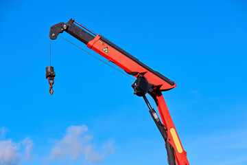 Red hydraulic loader crane boom with hook against a clear blue sky. Professional knuckle boom crane for industrial lifting, construction, and cargo handling. Heavy-duty truck-mounted equipment. © Igor