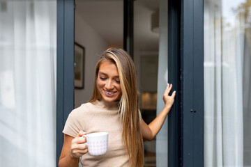 Fototapeta premium Smiling woman standing by balcony door with coffee cup. Morning relaxation, home comfort, and lifestyle concept. Happy female holding mug and looking down. Peaceful moment in modern apartment home.