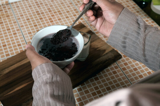 Woman's hand stirring a bowl of Bubur Ketan Hitam (black sticky rice), a sweet Indonesian porridge made from black sticky rice, coconut milk, and palm or cane sugar.  Indoensian traditional food. 