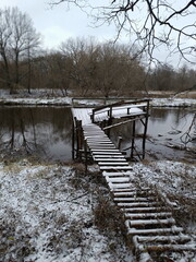 Fototapeta premium Rustic wooden pier on a river covered in first snow. Winter landscape with a jetty and bare trees. Cold, quiet scenery by the water
