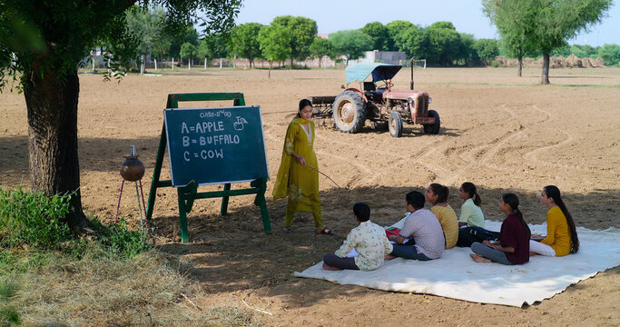 Indian Young adult gen z female tutor standing teach English to villager girl boy outdoor rural area. Happy child group sitting on mat look board learn do study new day time outside ground place