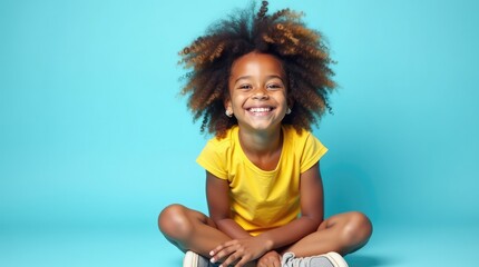 Smiling young girl with vibrant afro sitting cross legged on floor against bright blue background wearing yellow shirt