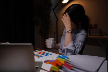 Thoughtful Asian businesswoman working late at night in dark office with laptop and paperwork