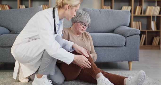 Doctor helps senior woman patient with injury. She assesses leg pain on the floor at home, offering careful help, comfort, examination and ongoing care. Compassionate medical support at home.