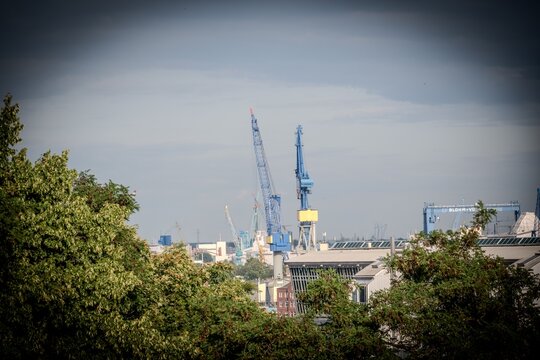 A crane construction site in Hamburg, Germany, with a cloudy sky and lush foliage.