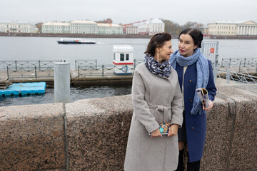 Obraz premium Two adult women standing near harbor waterfront discussing something while looking toward water and pier on cloudy day.