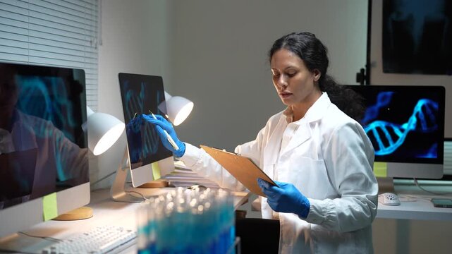 Genetic engineer in a white lab coat studying a dna helix on a computer screen and taking notes on a clipboard inside a laboratory with test tubes and x-rays on monitors in the background