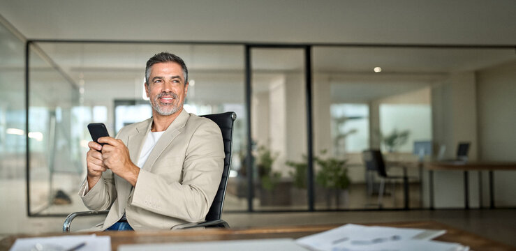 Happy busy middle aged business man investor wearing suit sitting in office using mobile cell phone. 50 years old businessman professional entrepreneur banker holding smartphone looking away at work.