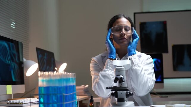 Focused female laboratory researcher in a white coat and gloves puts on safety glasses and begins examining a specimen with a microscope in a modern science facility at night