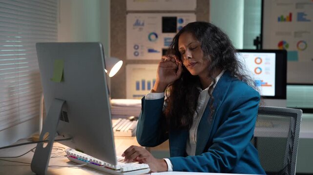 Exhausted and sleepy corporate worker feeling a headache while working late at night on a desktop computer, struggling to meet a deadline in a modern office with financial charts on the wall