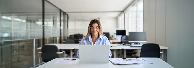 Happy smiling mature middle aged professional business woman investor manager executive or attorney looking at laptop at workplace working on computer in office.