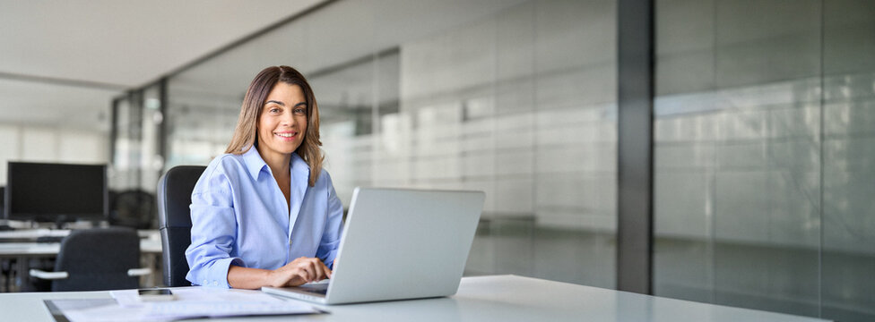Happy smiling mature middle aged professional business woman bank worker or hr manager, corporate executive or lawyer attorney looking at camera at work working on laptop computer in office, portrait.