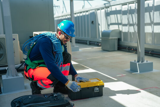 Professional HVAC technician wearing protective helmet and safety harness crouches on commercial building rooftop opening toolbox near air conditioner condenser units preparing repair equipment.