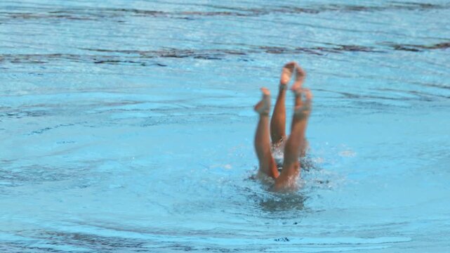 Two artistic swimmers performing synchronized leg movements during a duet routine in a swimming pool