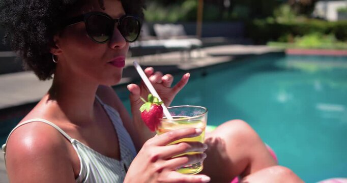African American woman seeing drink at pool reaching sipping fixing shades in striped top, cooling