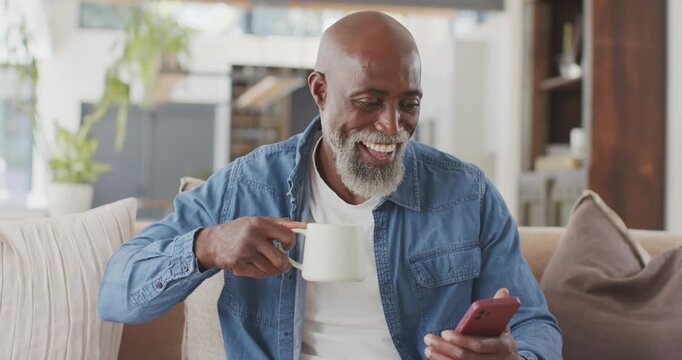 Mature man in denim shirt reading red smartphone, sipping mug on home couch, laughing at content