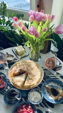 Luxury Maslenitsa breakfast: Russian crepes with black caviar, white wine and pink tulips. Elegant table setting with a black cat among houseplants on the background. Expensive lifestyle concept.