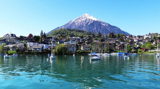 Spiez Schiffstation n snowcappep mountain with white snow peak or swiss alps over house village city on calm water wave with boat ship or cruise ferry on blue sky, 4k Lake Thun Interlaken Switzerland