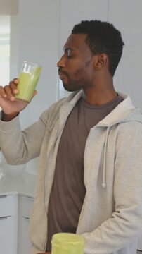Vertical video: Lifting glass man in gray hoodie sipping green smoothie at kitchen eyeing mixer cup