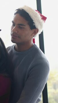 Vertical video: Leaning man hugging woman at window for holiday, wearing Santa hat and antlers