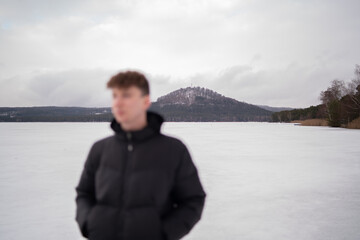 Fototapeta premium View of snowy hill across Macha Lake with blurred man in foreground. Winter landscape in Czech Republic. Man standing near frozen Macha Lake in Czech Republic looking at distant hill. Winter travel an