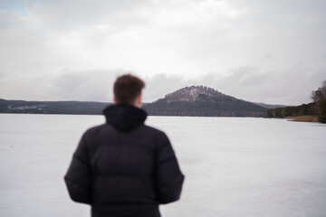 Fototapeta premium View of snowy hill across Macha Lake with blurred man in foreground. Winter landscape in Czech Republic. Man standing near frozen Macha Lake in Czech Republic looking at distant hill. Winter travel an
