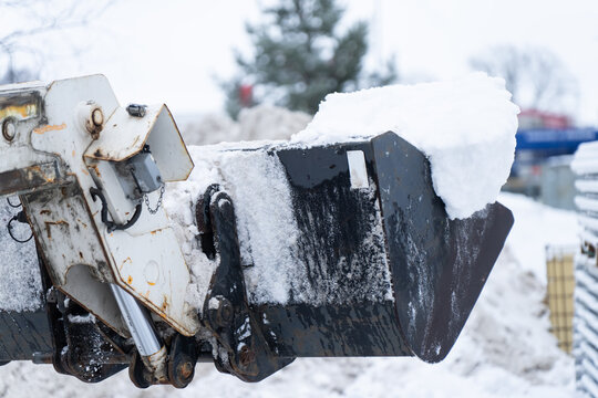 Industrial winter closeup of black digging bucket with thick snow cap and icy residue, hydraulic cylinders and pins visible, earthmoving equipment detail