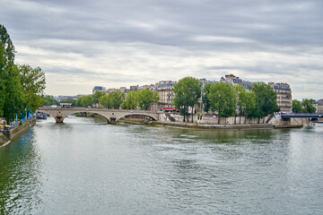 Obraz premium Scenic view of Seine River in Paris with historic stone bridge, Haussmann buildings, and tree-lined embankment under dramatic cloudy sky
