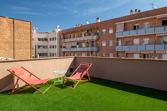 A sunny rooftop terrace in Barcelona with two red lounge chairs and a small table on artificial grass. Surrounding buildings are visible in the background.