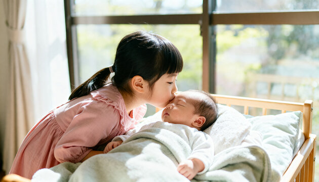 Little girl gently kissing sleeping newborn baby brother on forehead in crib at home. Concept of family, sisterhood, and new sibling love.