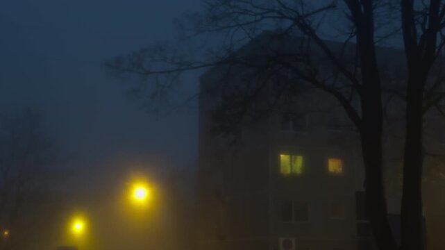 Foggy night scene. Misty evening reveals illuminated apartment facades. Dense fog outlines house silhouettes with glowing interior lights