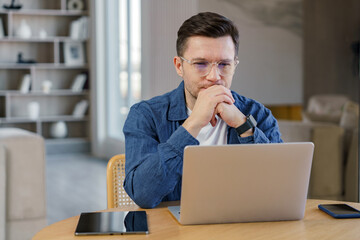 Man intently focused on laptop screen while working remotely from modern home office at a bright...