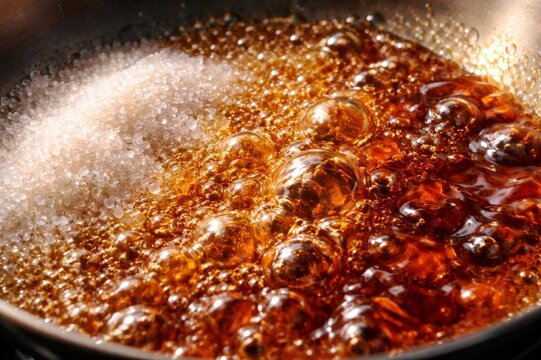 Sugar granules melting into a bubbling, glossy caramel in a hot saucepan, close up of homemade confectionery preparation showing sticky, golden brown texture and boiling motion