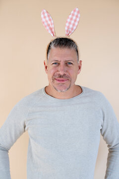 Man with grey hair and brown sweater with Easter bunny ears standing in front of brown background