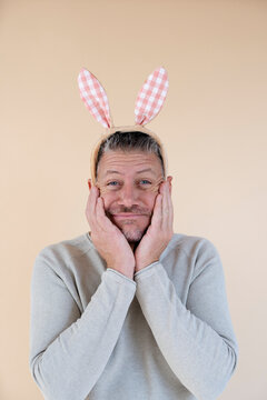 Man with grey hair and brown sweater with Easter bunny ears standing in front of brown background