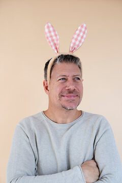 Man with grey hair and brown sweater with Easter bunny ears standing in front of brown background