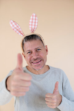 Man with grey hair and brown sweater with Easter bunny ears standing in front of brown background