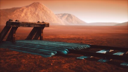 A wooden rail platform stands alone in a vast desert landscape, surrounded by distant mountains. The sky glows warmly, hinting at a unique sunset or sunrise moment. © icetray