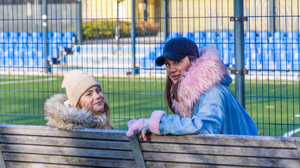 A smiling mother and her teenage daughter sitting together on a wooden park bench near a green soccer pitch with blue stadium seating, posing while waiting for a game on a sunny autumn day © Igor